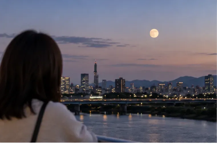 夕暮れの都市と川の向こうに昇る明るい月を眺める人