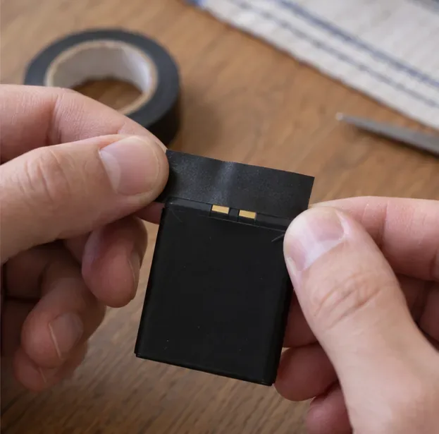 Due mani tengono una batteria integra con i contatti metallici esposti e una striscia di nastro isolante pronta per la preparazione al conferimento.