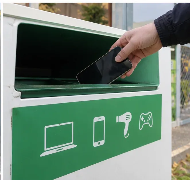 Une main dépose un smartphone entier dans un conteneur vert destiné aux appareils électriques et électroniques usagés.