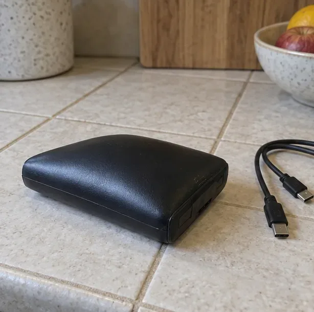 Une batterie externe noire posée sur un carrelage, avec ses câbles débranchés à côté.