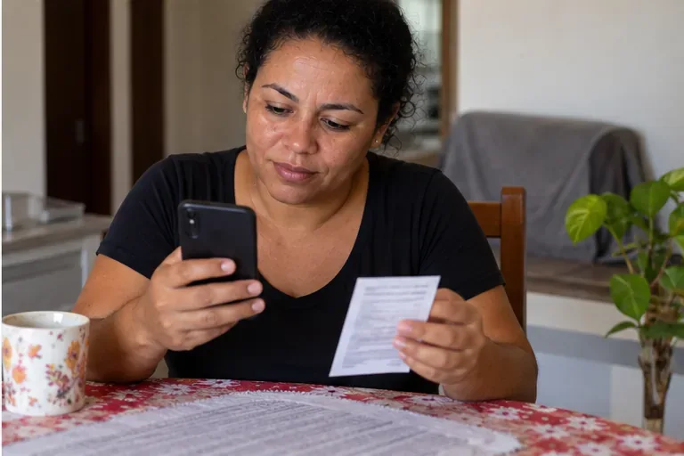 Mulher em casa conferindo o celular e um comprovante de atendimento depois de tratar uma pendência do Bolsa Família
