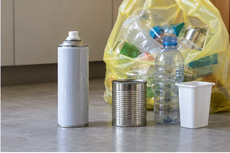 Una bomboletta spray integra è appoggiata accanto a un sacco giallo trasparente con altri imballaggi di plastica e metallo.