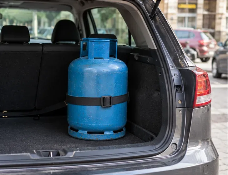 Une bouteille de gaz bleue est maintenue debout par une sangle dans le coffre ouvert d’une voiture.