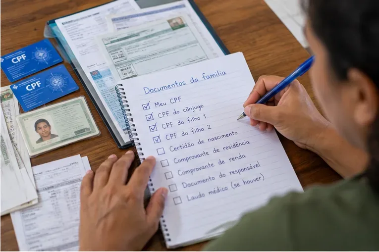 Pessoa organiza uma lista de documentos da família ao lado de CPFs e documentos de identificação.