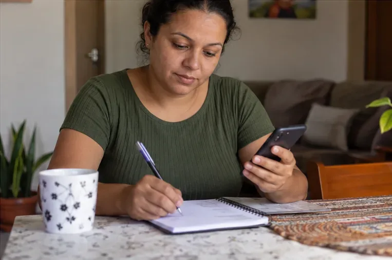 Pessoa em casa consultando o celular enquanto anota informações em um caderno.