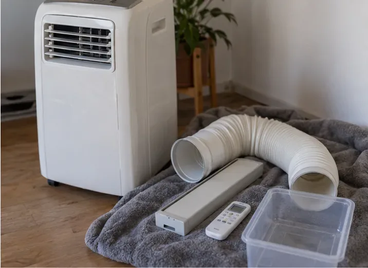 Un climatiseur mobile accompagné de son tuyau d’évacuation et de sa télécommande posés à côté.