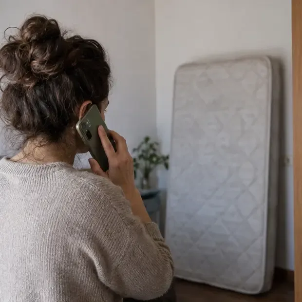 Una donna telefona con lo smartphone mentre un materasso usato è appoggiato in sicurezza vicino a una parete