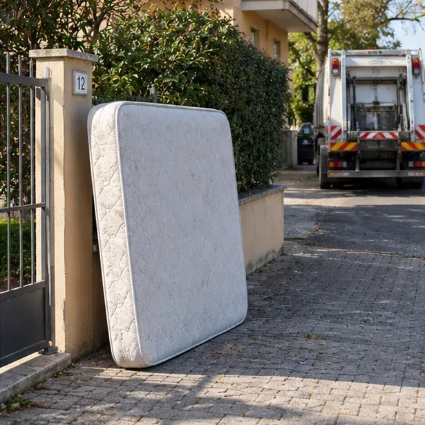 Materasso appoggiato vicino a un ingresso privato mentre un mezzo di raccolta si avvicina lungo la strada