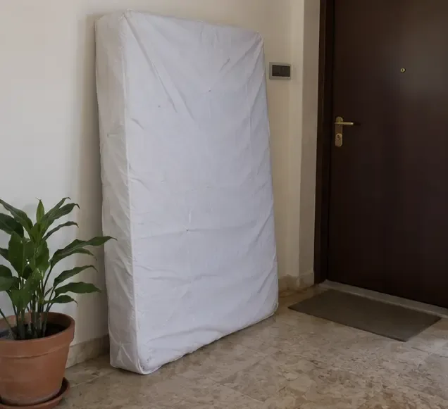 Un materasso protetto da una copertura chiara è appoggiato all’interno vicino all’ingresso