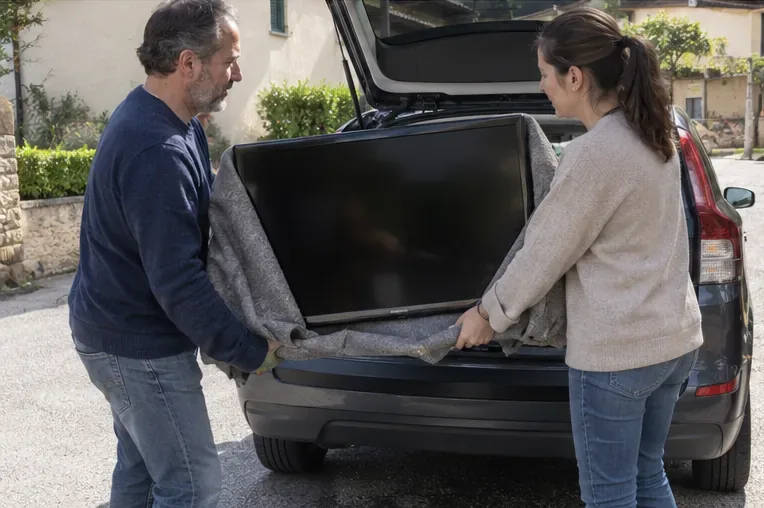Due persone trasportano in posizione verticale un televisore protetto da una coperta verso il bagagliaio di un’auto