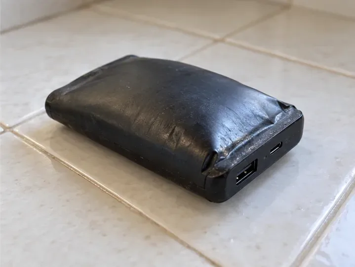 Un power bank con involucro deformato è appoggiato da solo su una superficie ceramica, senza essere maneggiato.