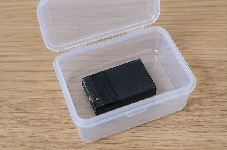 Piccola batteria al litio con terminali protetti posata da sola in un contenitore di plastica trasparente