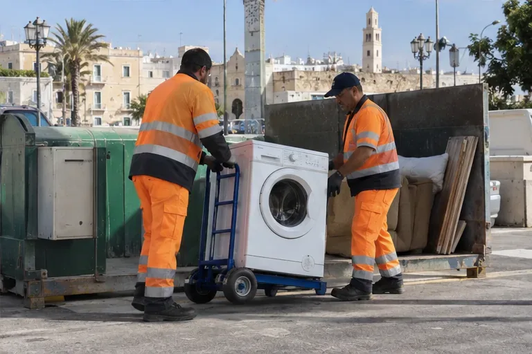 Due operatori in abbigliamento ad alta visibilità spostano una lavatrice su un carrello manuale presso un punto di raccolta