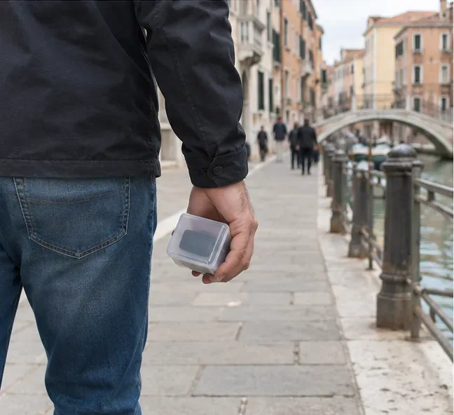 Una persona cammina lungo un canale veneziano portando una piccola power bank in un contenitore rigido.