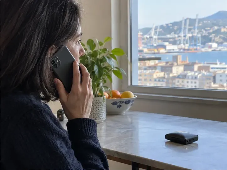 Persona al telefono davanti a una power bank gonfia appoggiata lontano su un tavolo, con il porto di Genova visibile dalla finestra