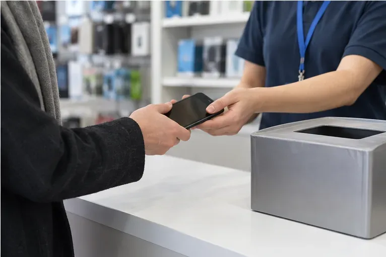 Un cliente consegna uno smartphone a un addetto in un negozio di elettronica vicino a un raccoglitore per piccoli dispositivi