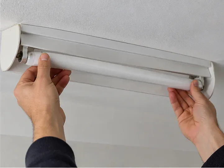 Deux mains retirent délicatement un tube fluorescent blanc d’un luminaire fixé au plafond, sans le plier ni le heurter.