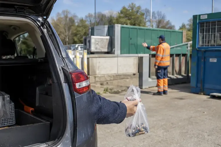 Une personne tient un petit sac de piles près du coffre ouvert d’une voiture, devant un centre de recyclage municipal où un agent l’oriente
