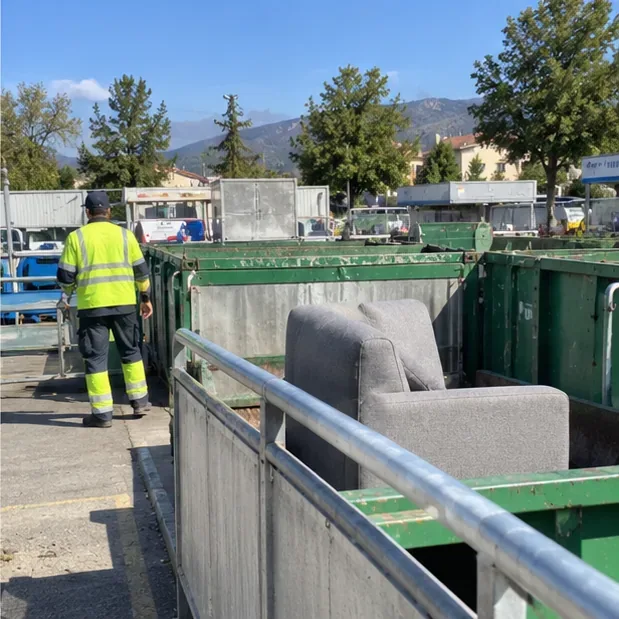Un canapé gris déposé dans une zone de collecte d’une déchèterie avec des bennes et un agent en tenue haute visibilité