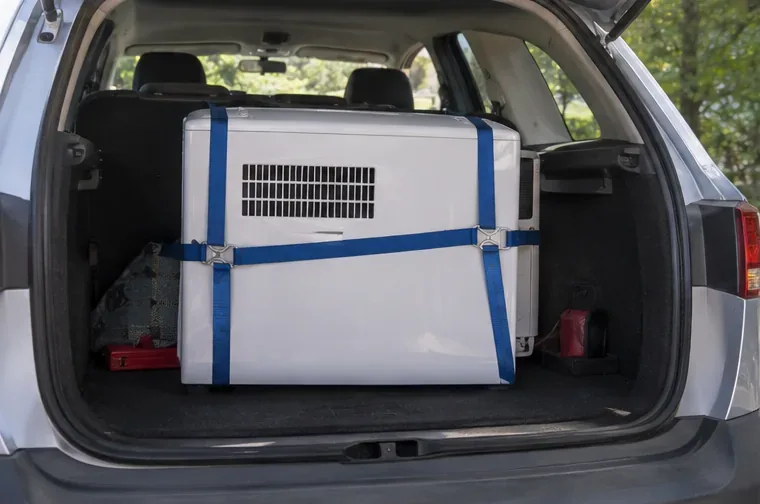 Climatiseur mobile blanc maintenu verticalement par des sangles dans le coffre d’une voiture