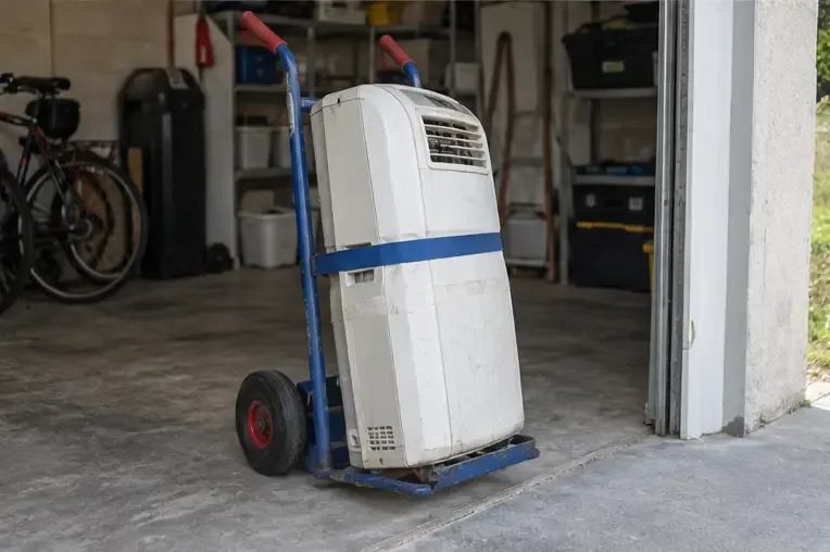 Climatiseur mobile usagé maintenu debout avec une sangle sur un diable dans un garage