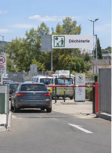 Une voiture entre dans une déchèterie avec une zone d’accueil et des conteneurs de tri visibles.