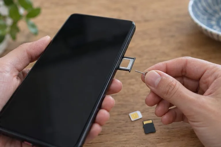 Gros plan sur les mains retirant le tiroir SIM d’un smartphone éteint avec une pointe d’éjection, tandis qu’une carte mémoire est posée sur la table.