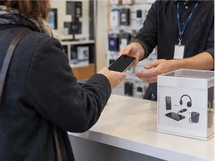 Une cliente remet un ancien smartphone à un employé au comptoir d’un magasin d’électronique.
