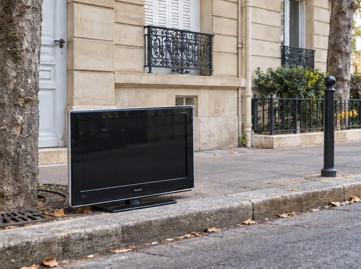 Un téléviseur à écran plat est posé devant un immeuble, près d’une entrée, sans autres déchets autour.