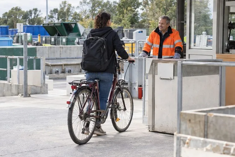 Une personne pousse un vélo usagé vers l’accueil d’une déchèterie et échange avec un agent en gilet orange.