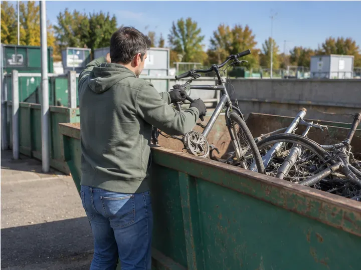 Un particulier dépose un vélo très endommagé dans une benne réservée aux métaux dans une déchèterie.