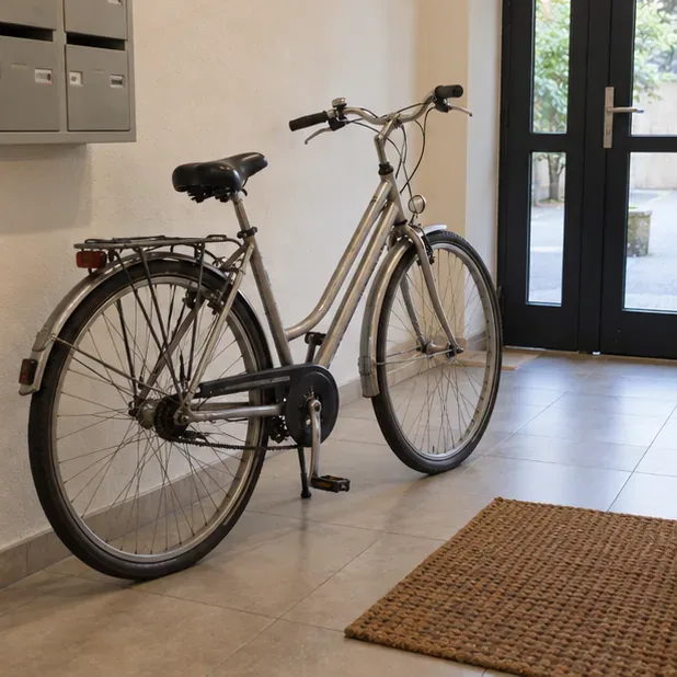 Un vélo est placé dans une entrée intérieure dégagée et éclairée, près d’une porte vitrée