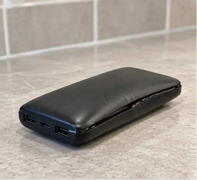 Batterie externe noire visiblement gonflée et déformée, posée seule sur une surface minérale