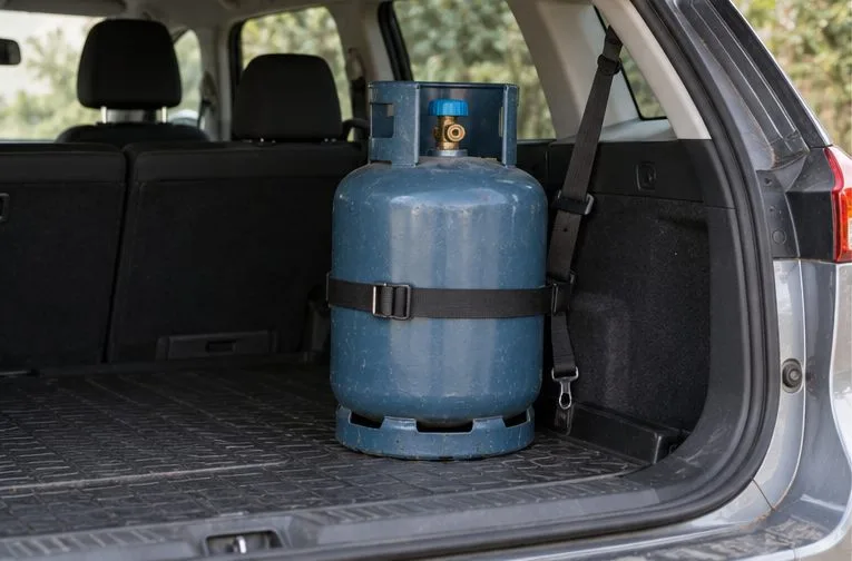 Une bouteille de gaz est maintenue debout par une sangle dans le coffre ouvert d’une voiture arrêtée.