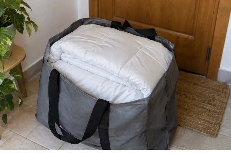 Une couette blanche propre et pliée est placée dans un grand sac de transport réutilisable près d’une porte d’entrée.