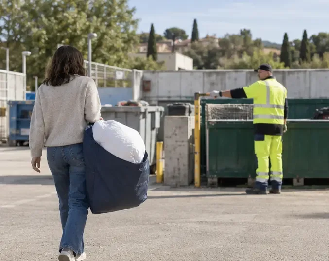 Une personne apporte une couette pliée dans une déchèterie tandis qu’un agent l’oriente vers la zone de dépôt