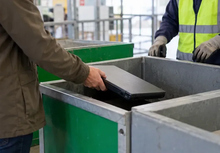 Une personne pose un ordinateur portable fermé dans un bac de collecte, sous le regard d’un agent en gilet fluorescent.
