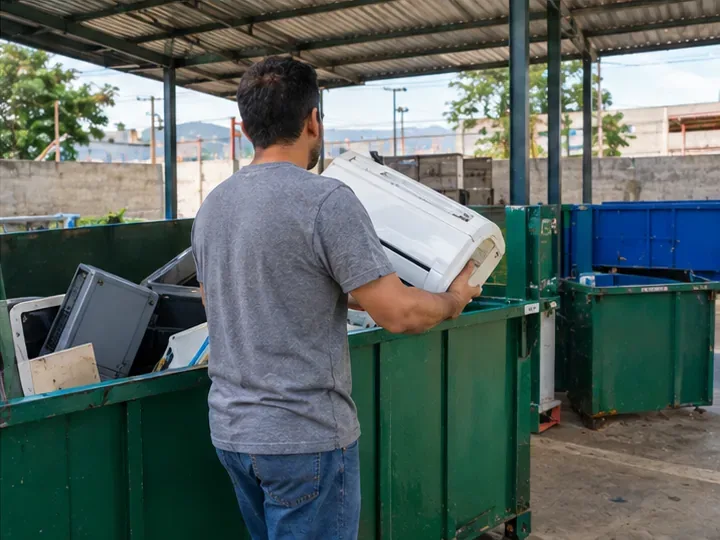 Homem entrega uma unidade de ar-condicionado em uma área de triagem com contentores para equipamentos descartados