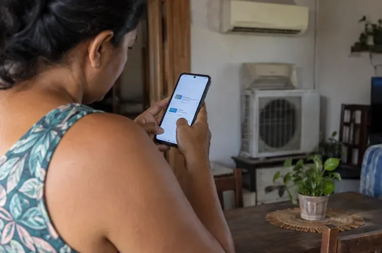 Moradora consulta no celular informações de coleta enquanto um ar-condicionado antigo permanece separado dentro de casa