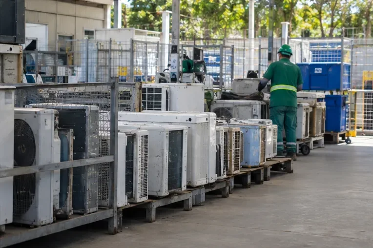 Área de triagem com várias unidades condensadoras de ar-condicionado organizadas sobre pallets em instalação de reciclagem.