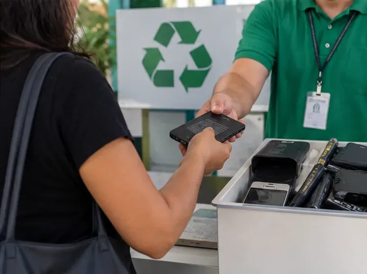 Pessoa entrega um equipamento com bateria em um balcão de reciclagem de eletrônicos, diante de uma caixa com aparelhos usados.