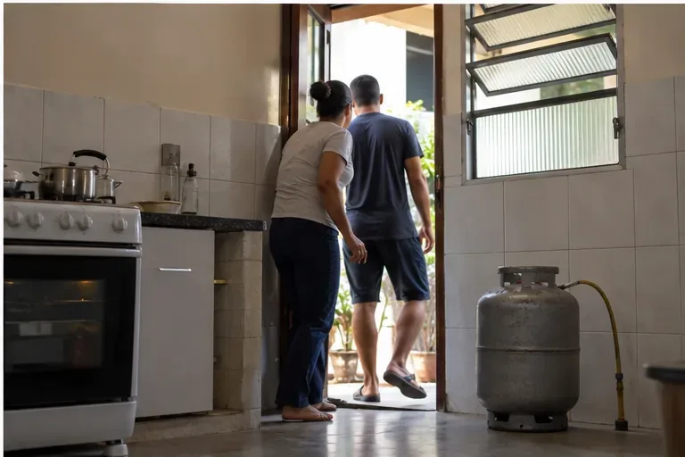Cozinha com porta e janela abertas enquanto duas pessoas deixam o ambiente e um botijão permanece afastado do fogão