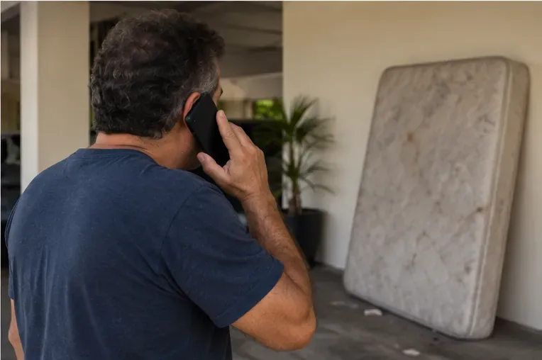 Homem usando o celular ao lado de um colchão encostado em área coberta