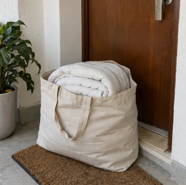 Um edredom branco limpo e dobrado está acomodado em uma bolsa de tecido reutilizável junto a uma porta