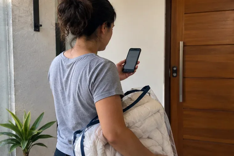Moradora segurando um edredom embalado enquanto consulta o celular junto à porta de casa.