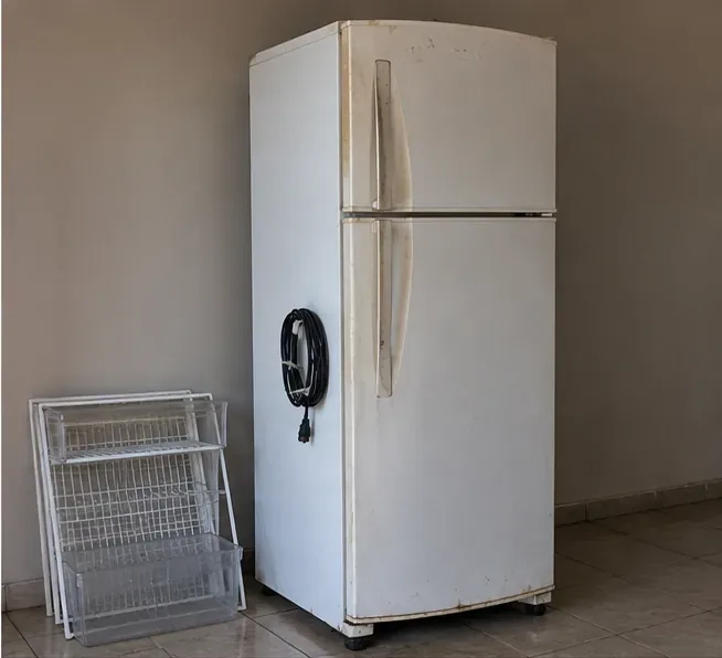Geladeira antiga fechada, com cabo preso na lateral e prateleiras removidas apoiadas ao lado