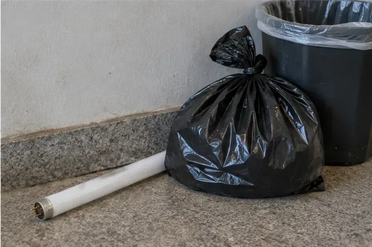 Tubo fluorescente deixado ao lado de um saco preto e de uma lixeira comum, exemplo de descarte inadequado