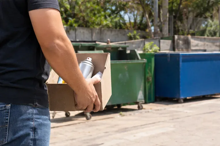 Pessoa levando uma caixa com materiais recicláveis e uma lata de spray para área de coleta com contêineres