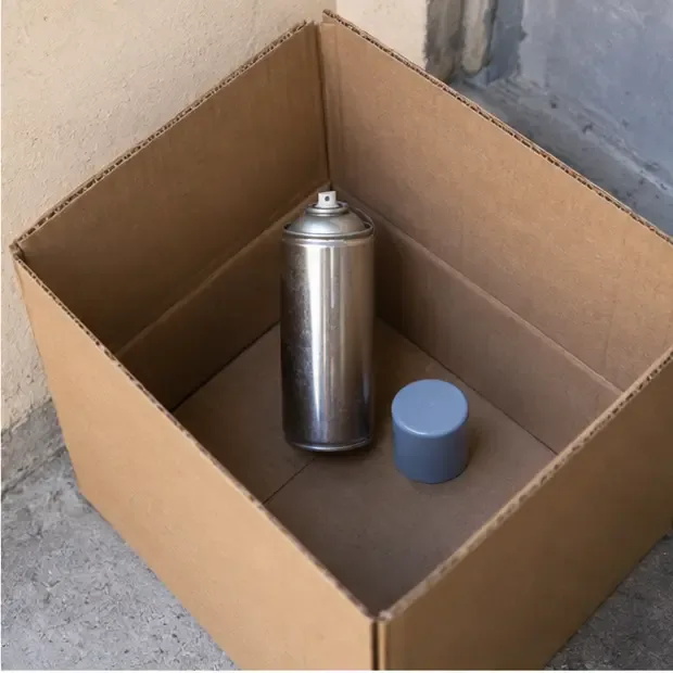 Lata de spray vazia com a tampa ao lado, separada dentro de uma caixa de papelão rígida