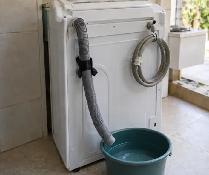 Máquina de lavar com mangueiras desconectadas e presas, enquanto a água restante escorre para um balde.